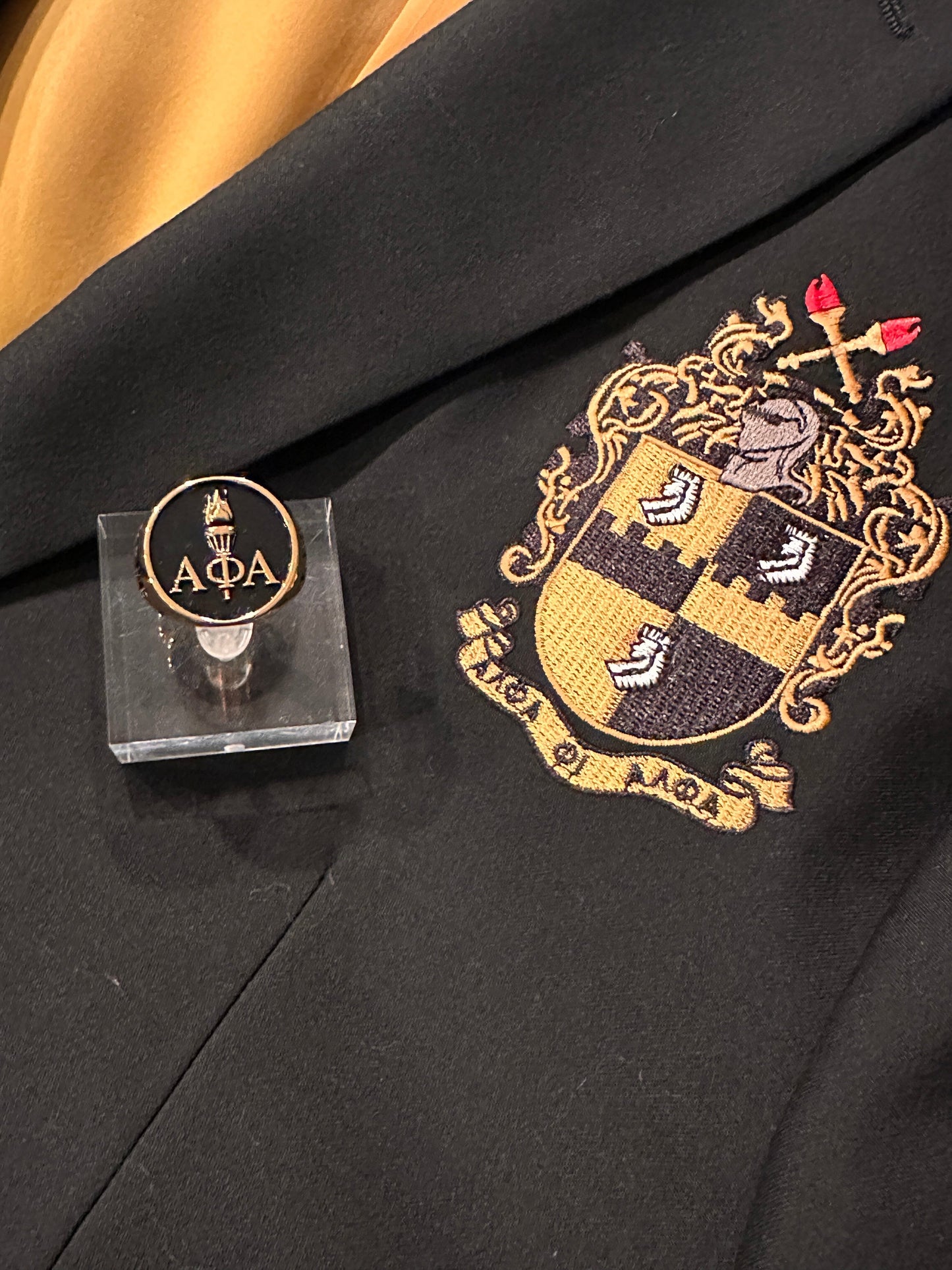 Gold Alpha Phi Alpha Light of the World ring with intricate A Phi A Greek letters and torch design, displayed on a black blazer with the fraternity's embroidered crest.