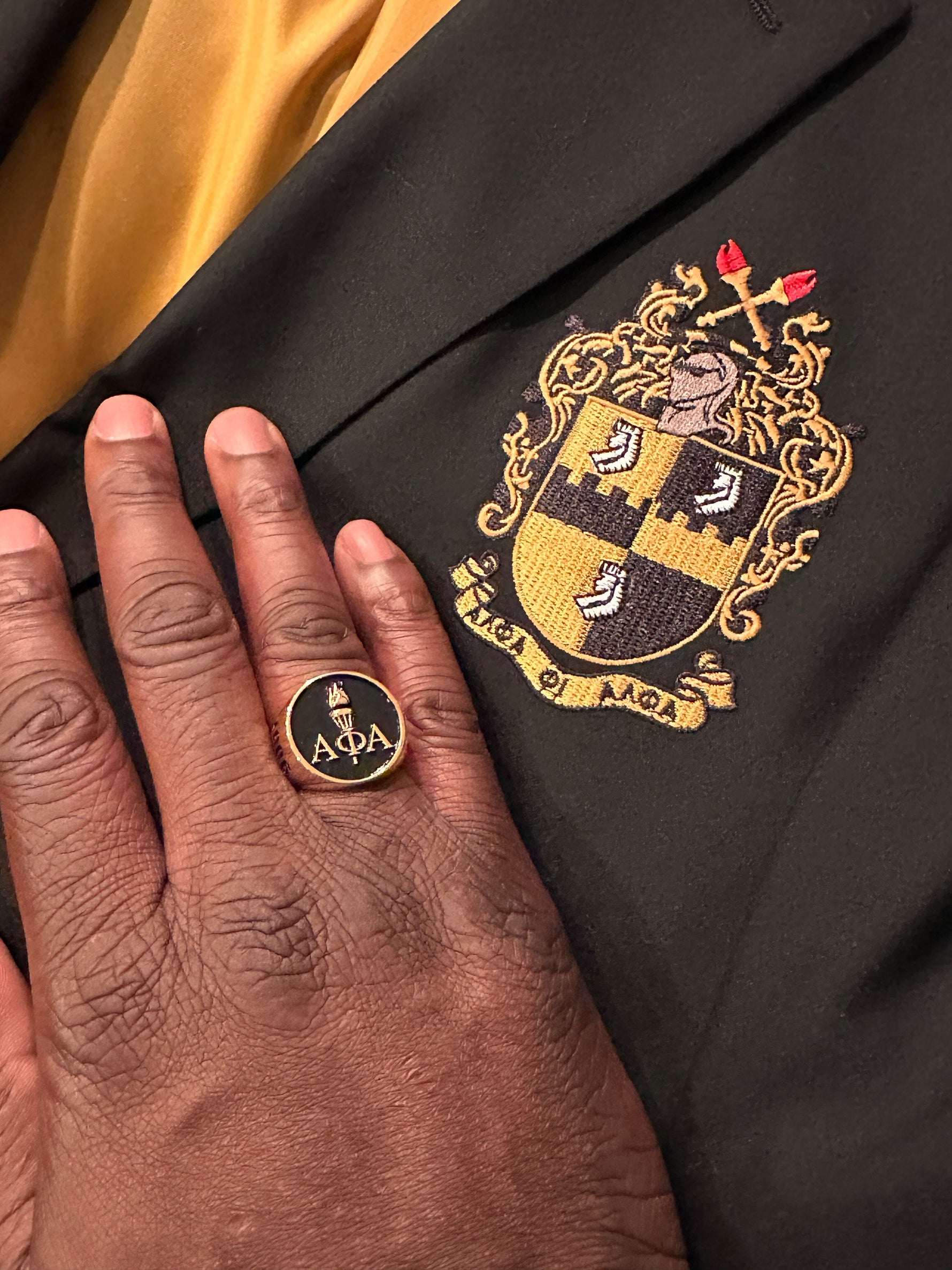 Alpha Phi Alpha Light of the World Gold Ring worn on a hand, displayed against a jacket with the Alpha Phi Alpha fraternity crest.