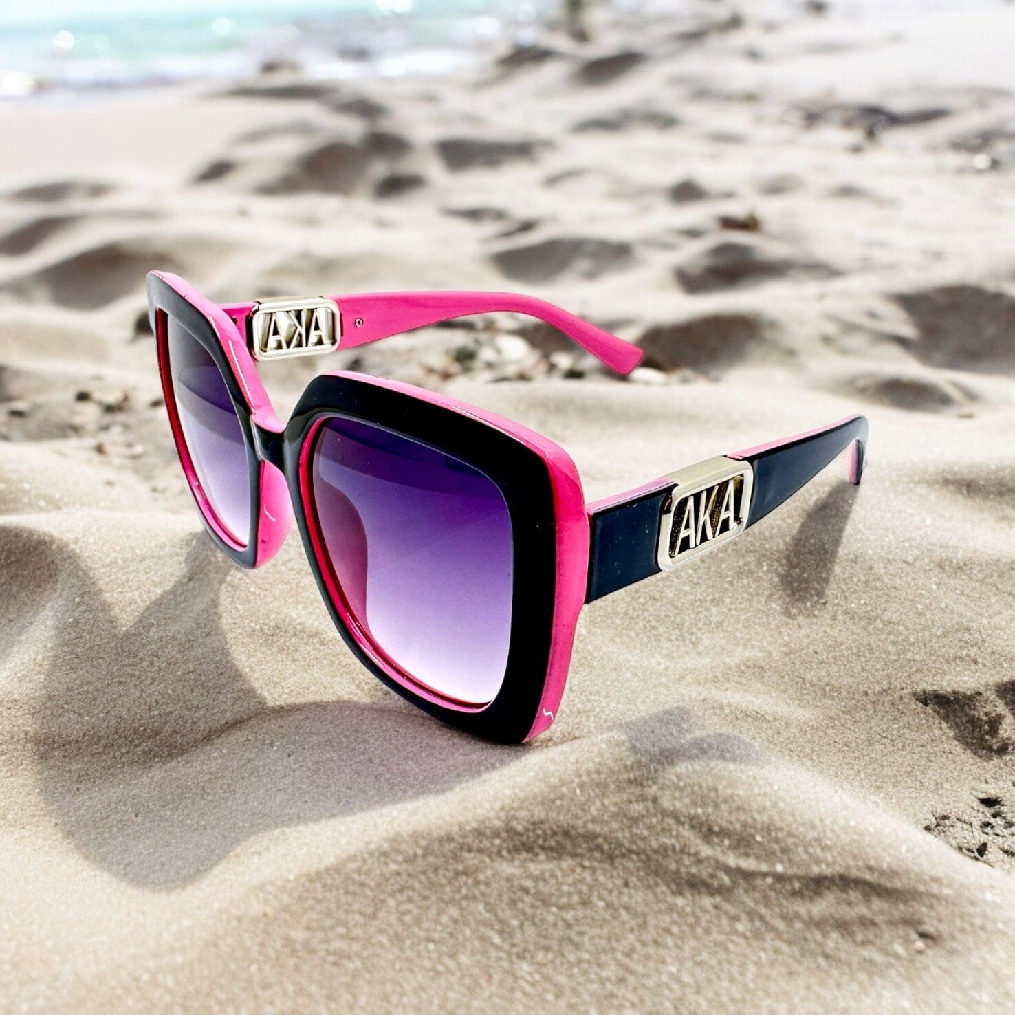 Elegant Pretty Girl Pink and Green AKA Sunglasses with UV protection and silver or gold AKA side letters, stylishly placed on sandy beach background.